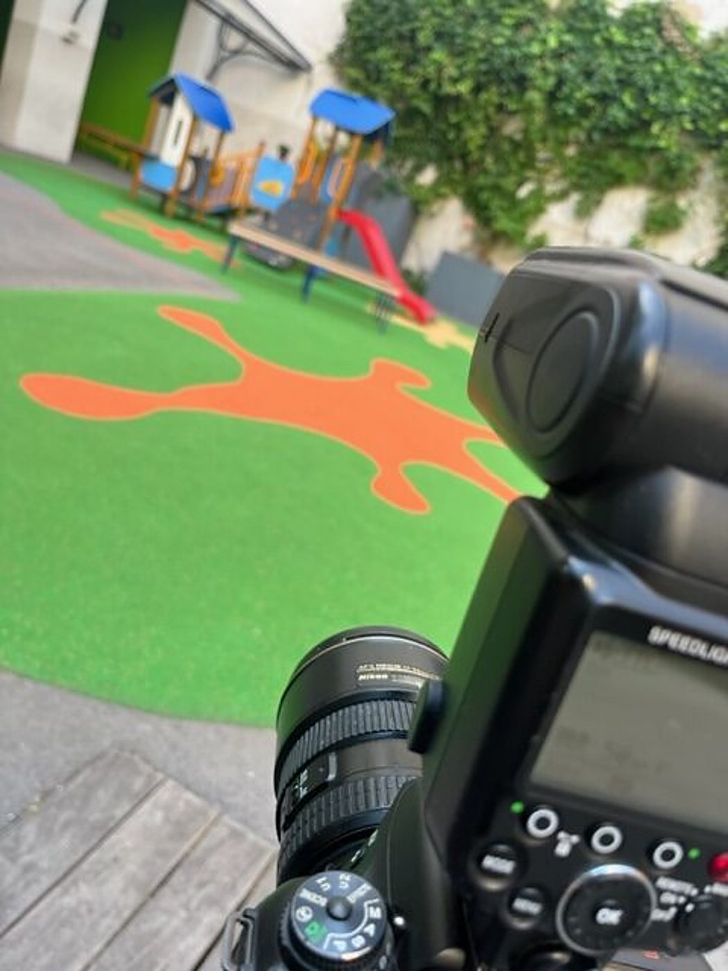Un appareil photo reflex avec flash au premier plan devant une aire de jeux pour enfants au sol de gazon synthétique vert et orange.