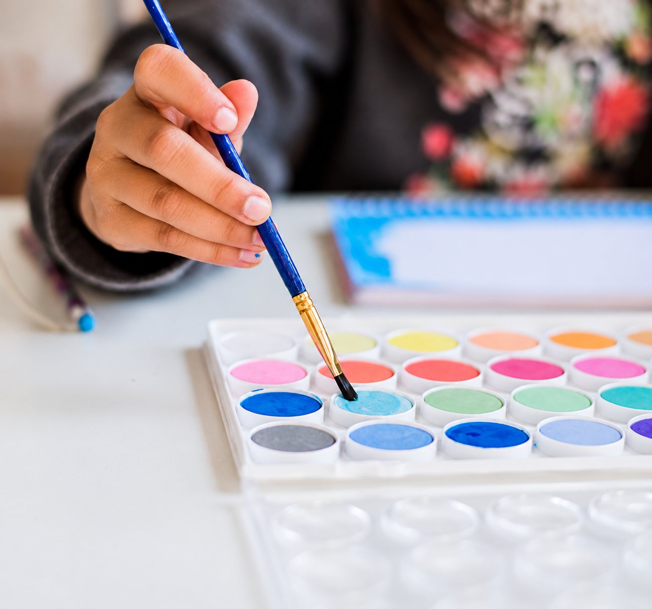 Une main tient un pinceau à manche bleu au-dessus d'une palette d'aquarelle aux godets de couleurs vives, posée sur une table blanche.