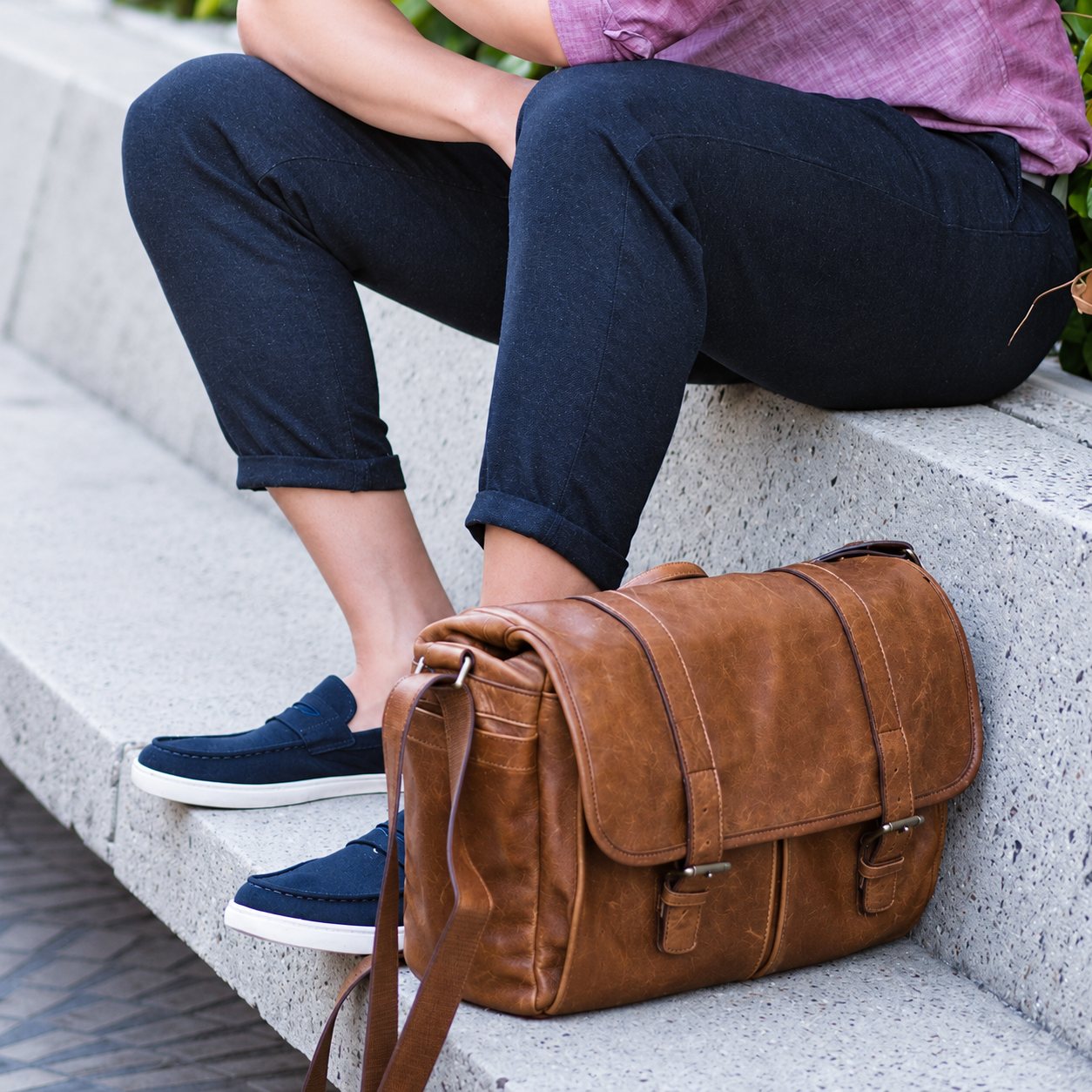 Un sac messager en cuir marron est posé sur un banc en béton, à côté des jambes d'un homme portant un pantalon sombre et des mocassins bleu marine.